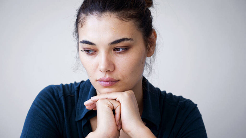 A woman with a forlorn expression rests her head on her hands and looks away from the camera.