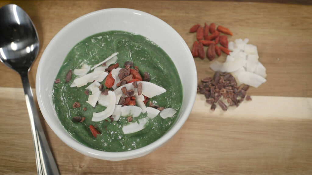 A smoothie bowl is pictured alongside a big spoon and toppings on a table.